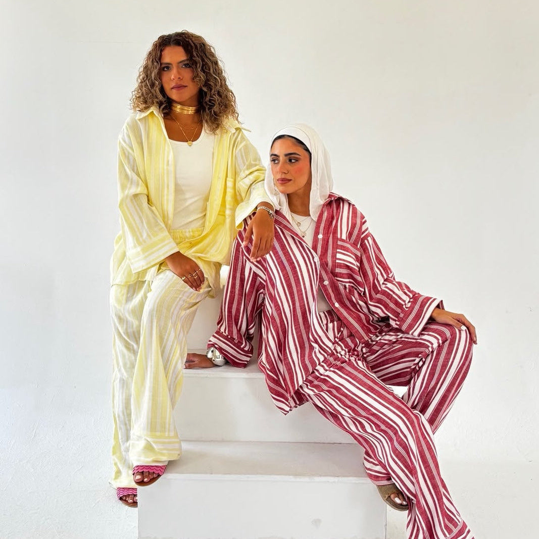 Two women modeling matching lounge sets in a bright studio. One wears a pale yellow robe and pants over a white top, while the other wears a red-and-white striped robe and pants with a white headscarf.